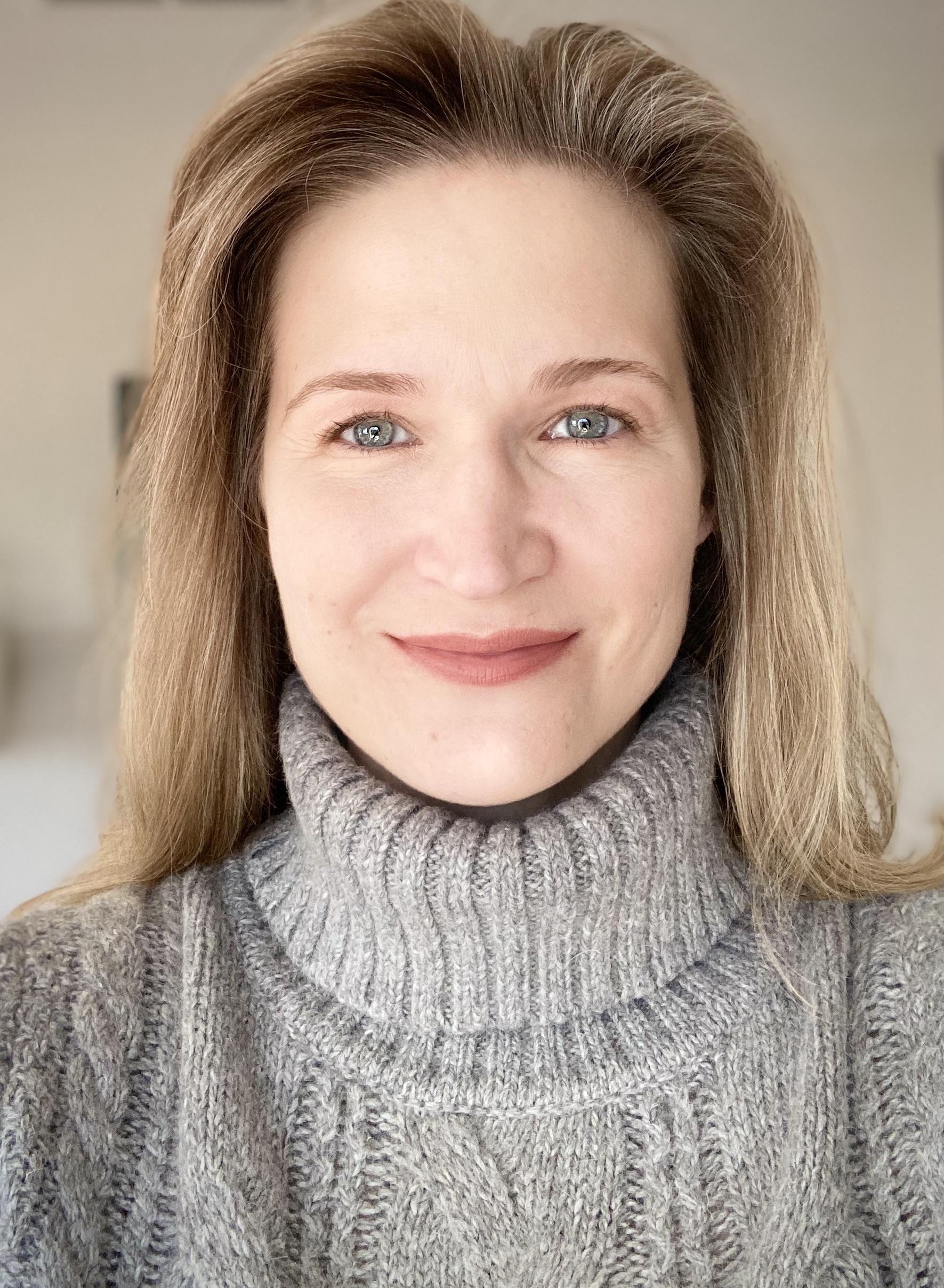 a blonde woman wearing a grey turtleneck sweater is looking at the camera